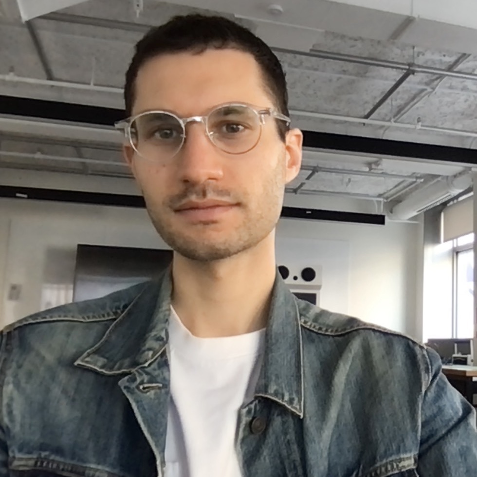 Skinny Jewish dude with short curly hair, clear glasses with a white shirt and denim jacket.